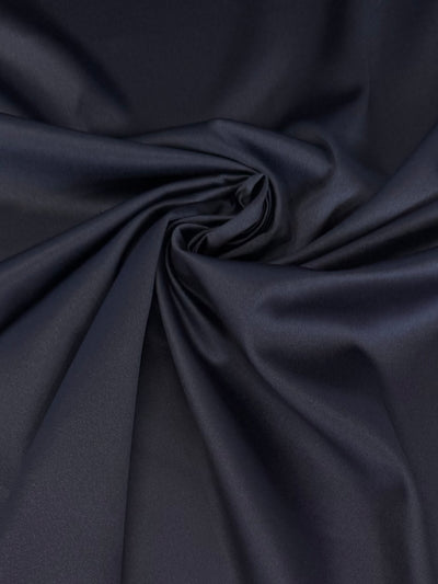 Close-up of Satin Deluxe - Navy by Super Cheap Fabrics, featuring a luxurious silky sheen reminiscent of nightgown satin. Gathered and twisted at the center, it creates soft folds and a swirl pattern, with its lustrous texture gently reflecting light across the surface.