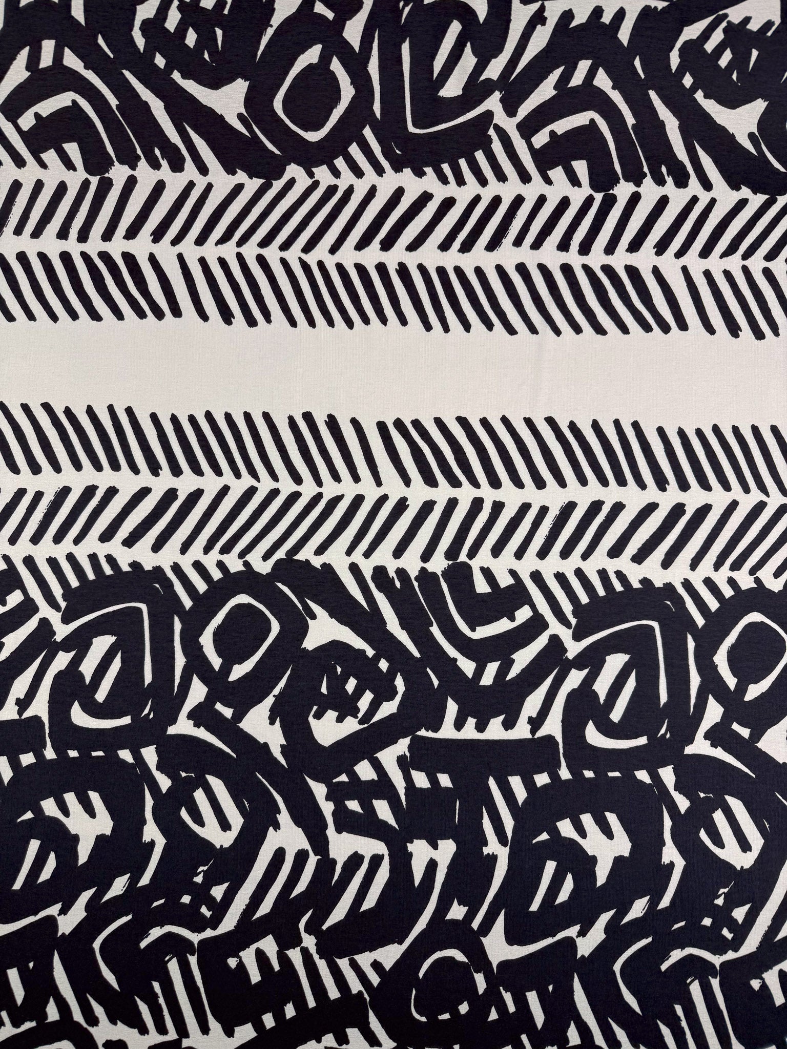 Super Cheap Fabrics’ Viscose Knit - Tribal Brushstroke (142cm) features abstract black shapes, parallel dashes, and bold organic patterns on beige—ideal inspiration for cool and breathable summer fabric designs.