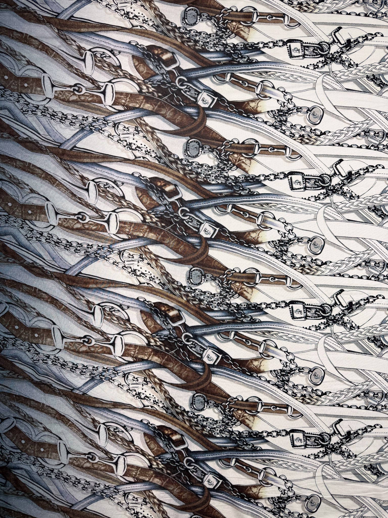 Super Cheap Fabrics' Viscose Knit - Link (138cm) features an intricate equestrian-themed pattern with brown leather bridles, bits, and chains overlapping on a white stretch fabric.