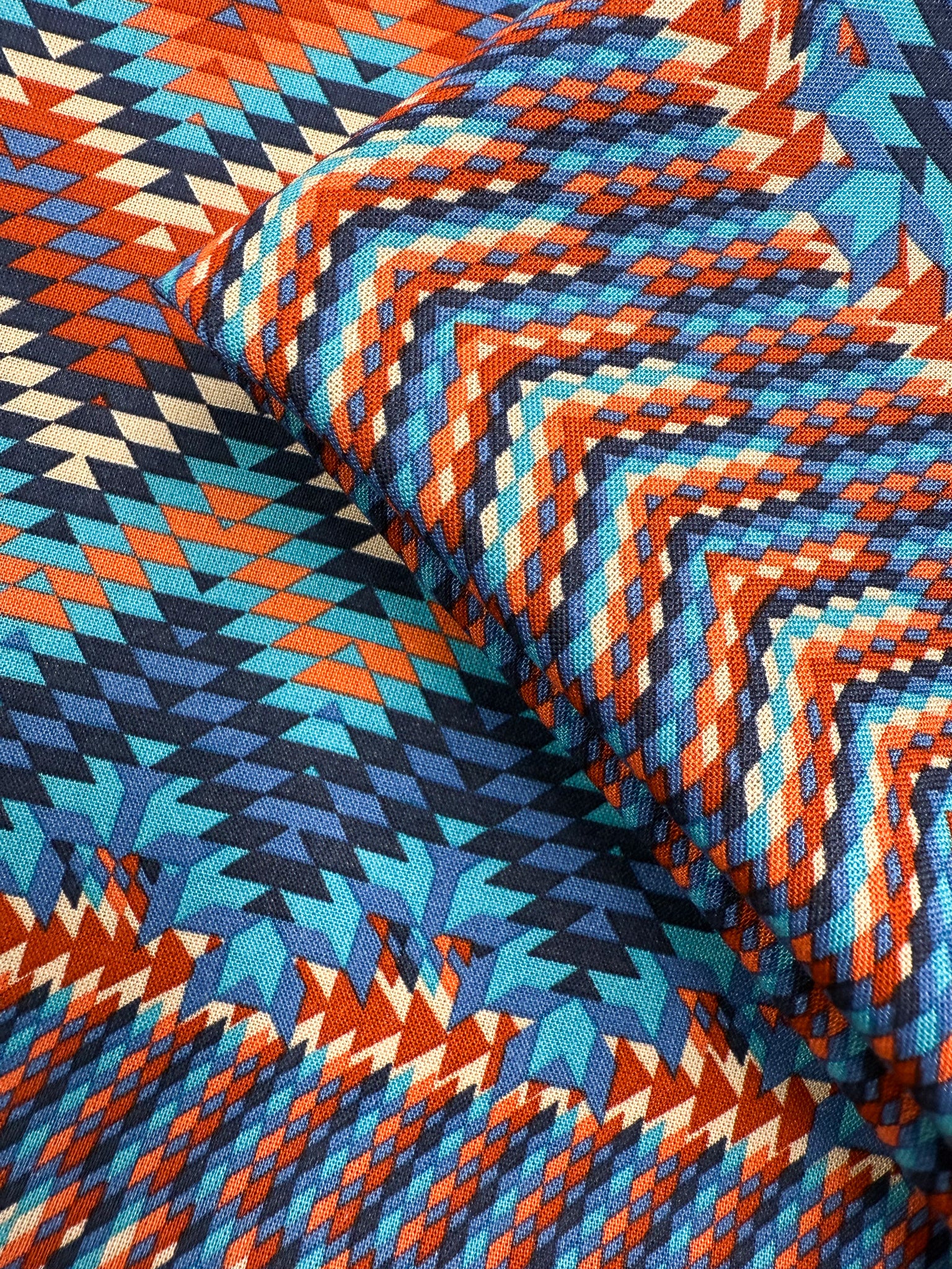 A close-up of Super Cheap Fabrics’ Printed Viscose - Zulu - 150cm shows geometric zigzag and diamond patterns in blue, orange, red, yellow, and beige from Prato, Italy. Slight folding adds depth and texture.