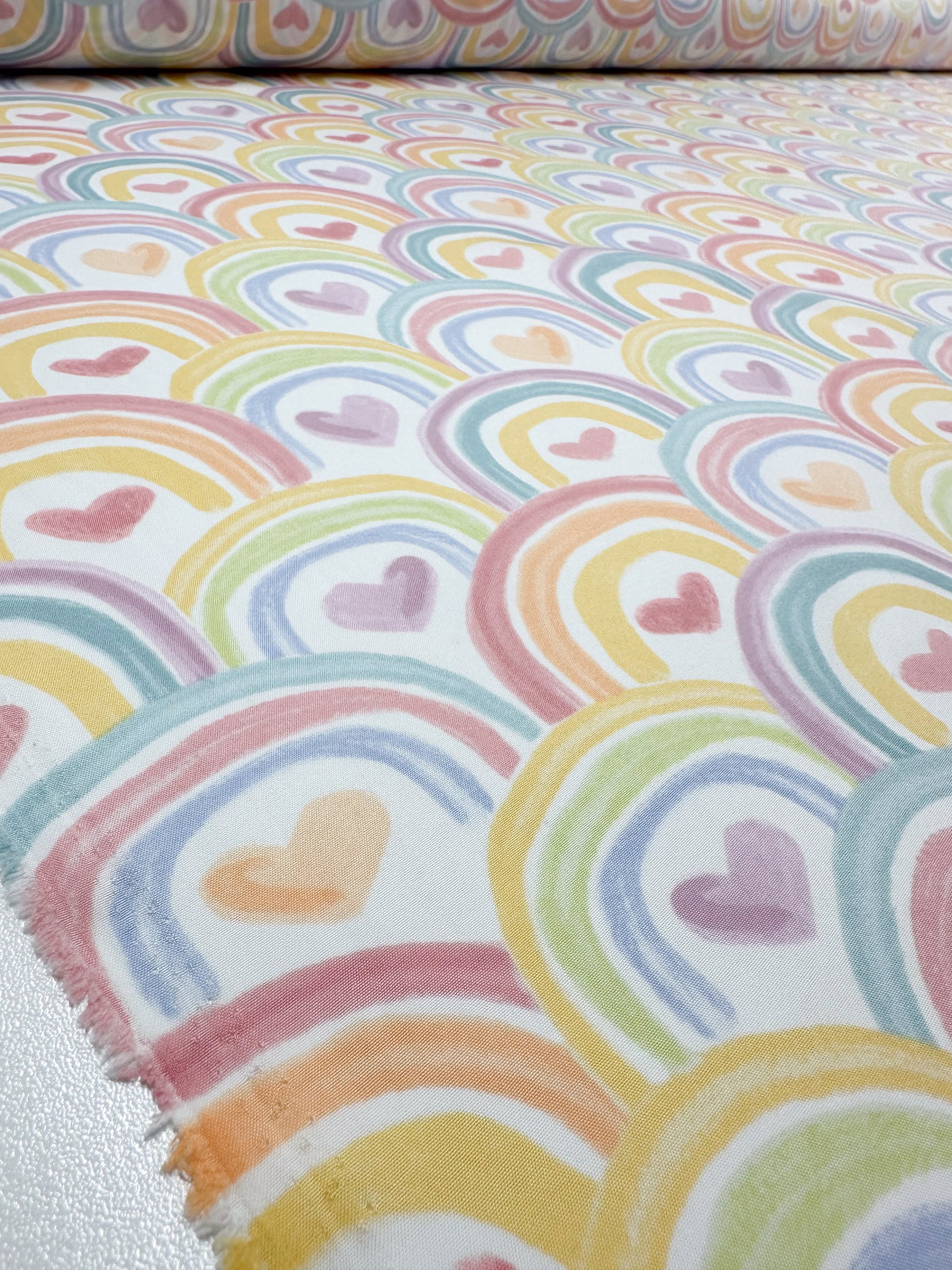 Close-up of the Unity Printed Delsutered Satin from Super Cheap Fabrics showcases overlapping rainbows with heart centers. The pastel hues in pink, orange, yellow, green, and blue form a whimsical design on this lightweight 155cm fabric reminiscent of printed satins.