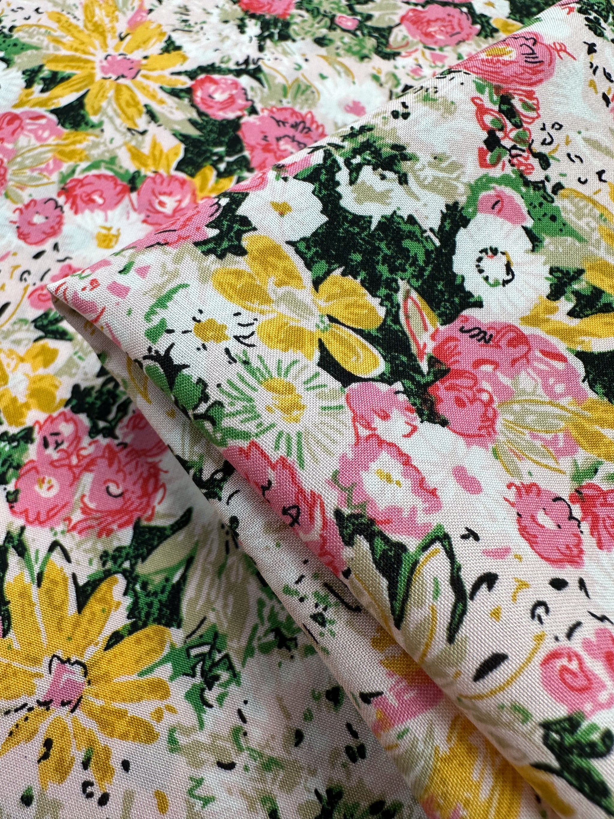 A close-up of Designer Rayon - Floral Garden by Super Cheap Fabrics shows vibrant yellow, pink, and white flowers with green leaves on a light background. Its lively print and soft drape make it a versatile 145cm-wide fabric for many projects.