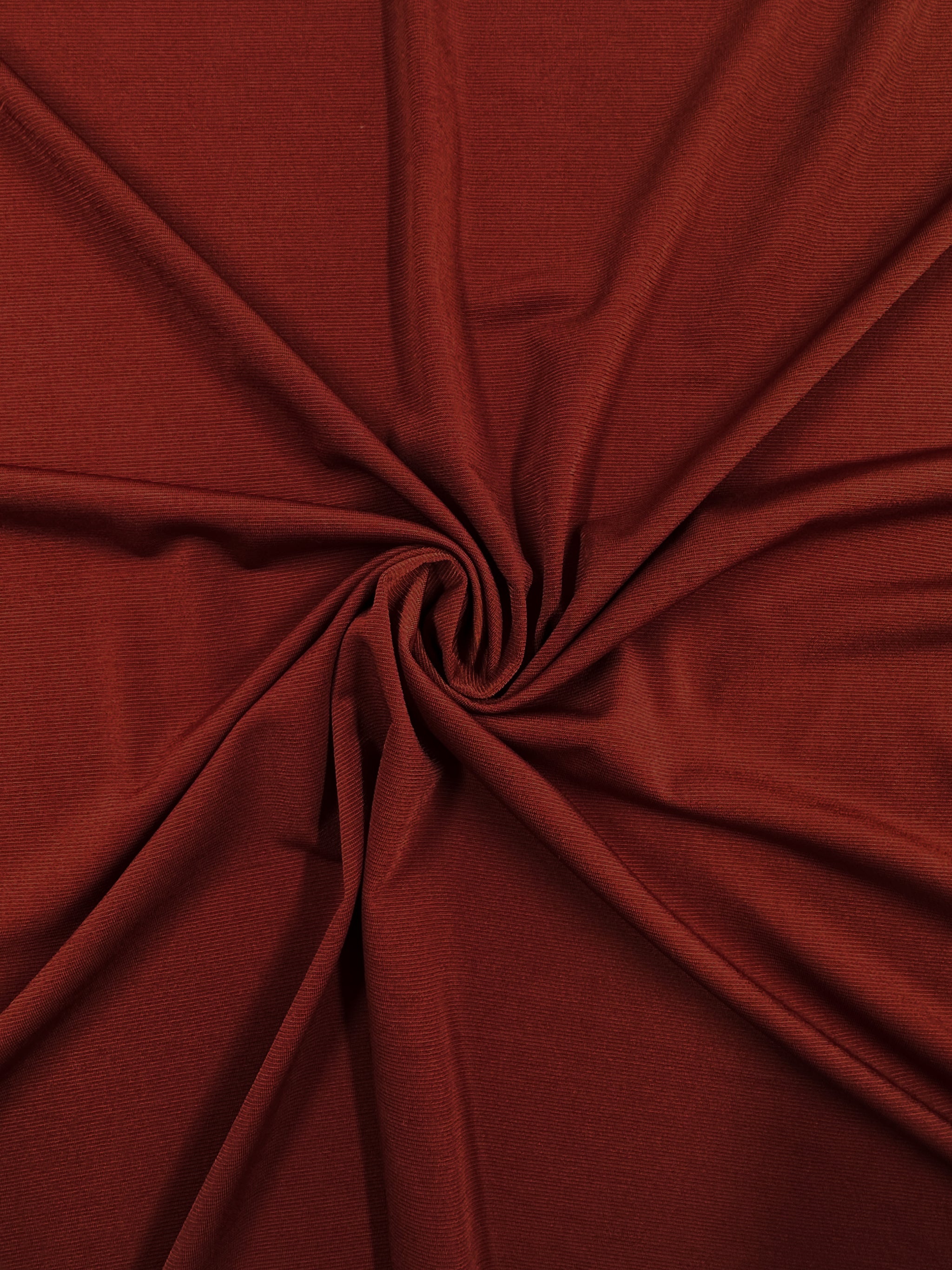 Close-up of Super Cheap Fabrics’ ITY Knit - Ketchup (150cm), a rust-colored polyester spandex with a soft texture, shown gathered and twisted at the center to create gentle folds and a circular pattern.