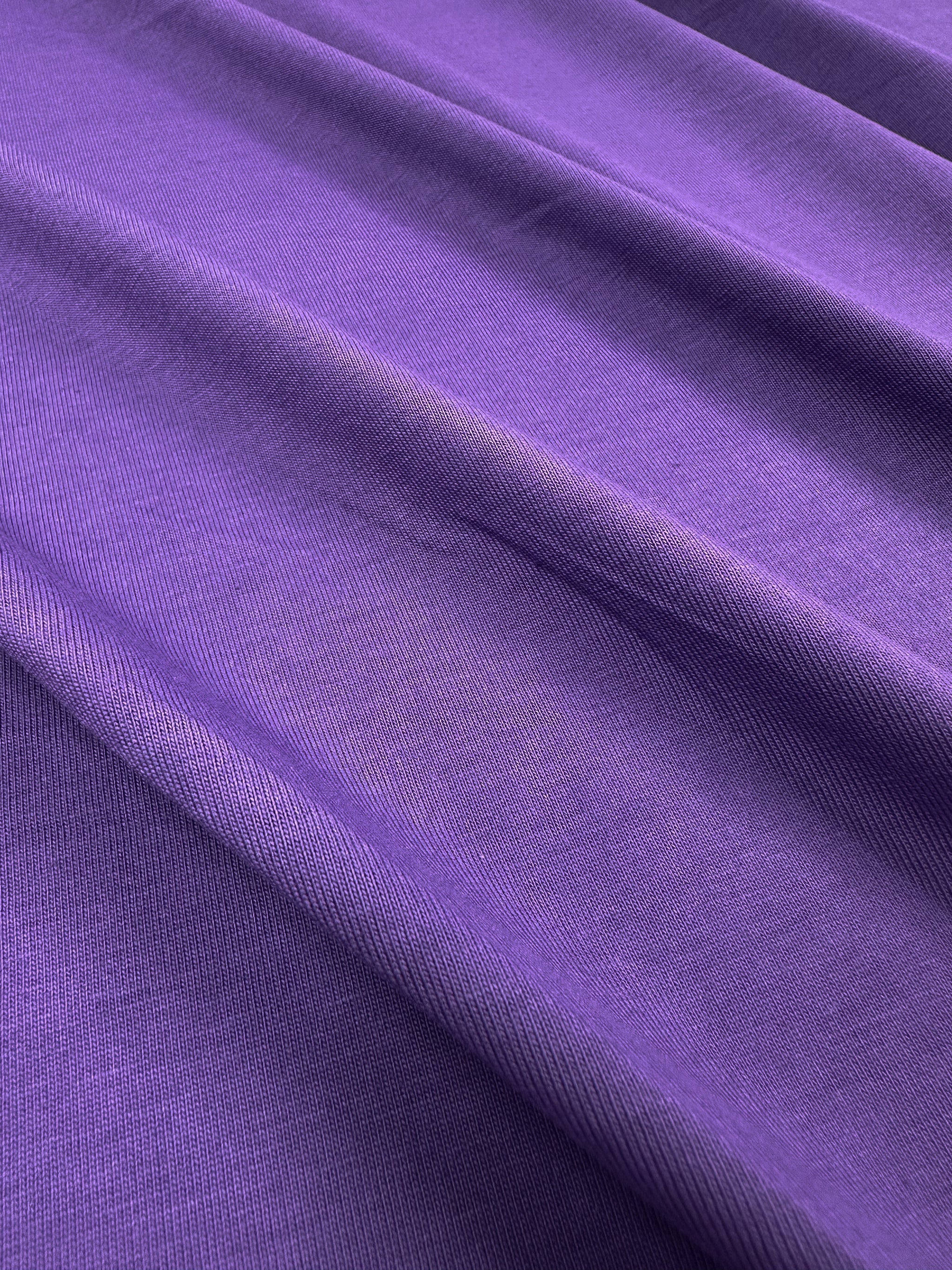A vibrant Patrician Purple cotton fabric from Super Cheap Fabrics drapes smoothly, highlighting its rich color and soft texture. The lighting enhances the elegance of this 170cm medium-weight Cotton Jersey, showcasing gentle folds.