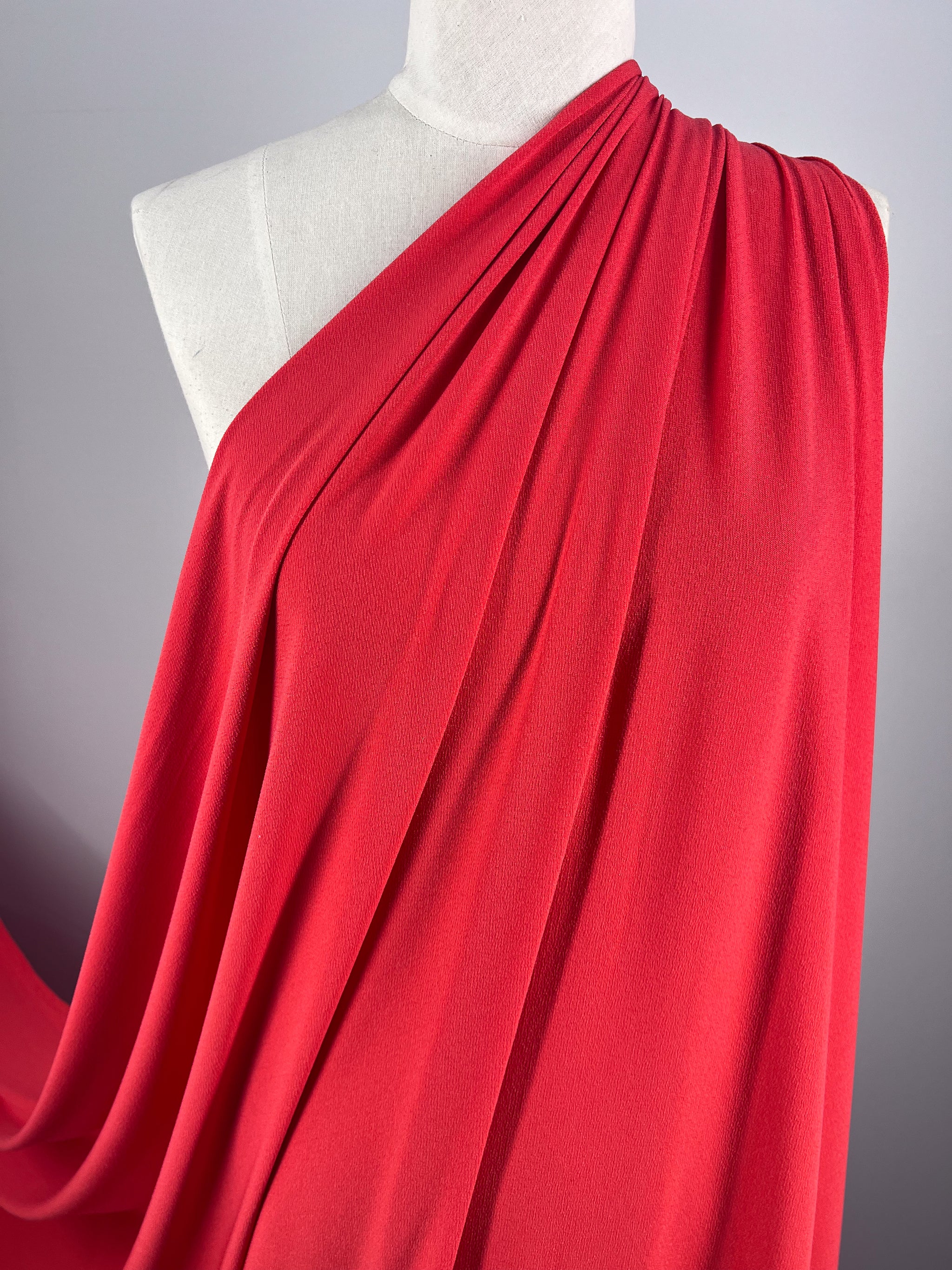 A mannequin draped in Super Cheap Fabrics' Crepe Lycra - Tangelo showcases vibrant coral pink fabric, gathered at the shoulder and elegantly cascading. The material's texture and pleats are highlighted against a neutral gray background.