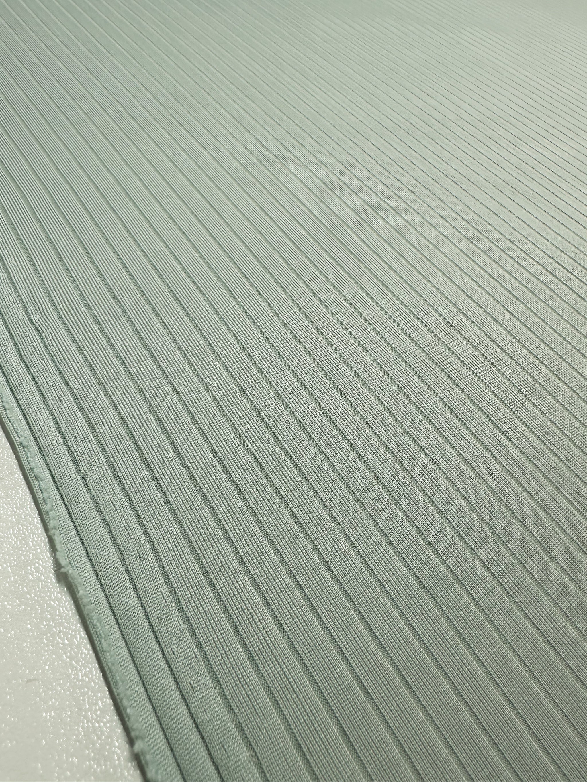 Close-up of Super Cheap Fabrics' Rib Knit - Murmur, 175cm: a medium weight, light green fabric with a smooth, uniform rib knit pattern. Subtle reflections enhance the linear design, while its generous stretch adds tactile appeal.