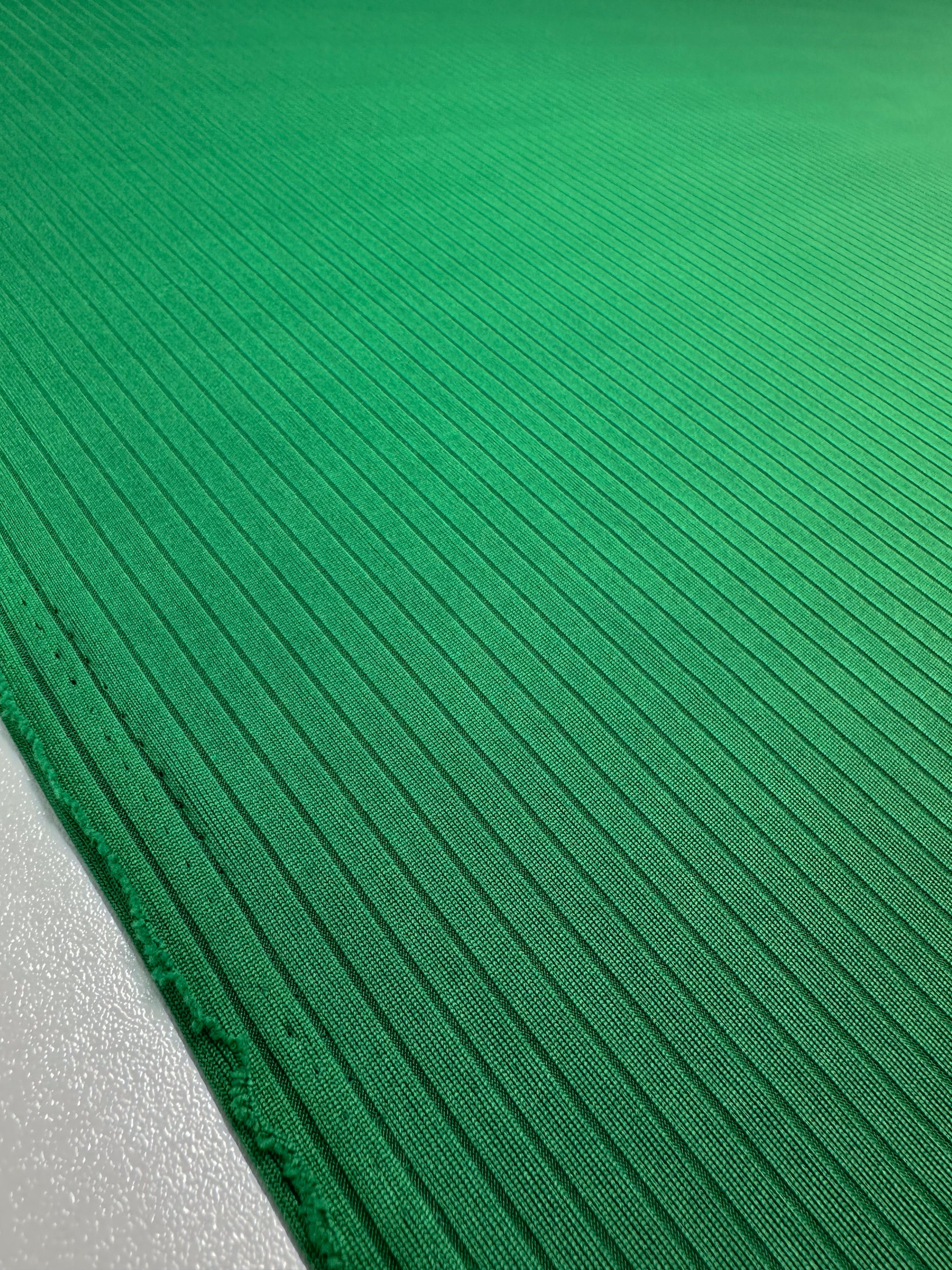 A close-up of the Rib Knit - Abundant Green fabric from Super Cheap Fabrics shows vibrant medium-weight green with diagonal thin ridges. The smooth and slightly shiny texture enhances its pattern, with the edge visible on the left side. Width: 177cm.