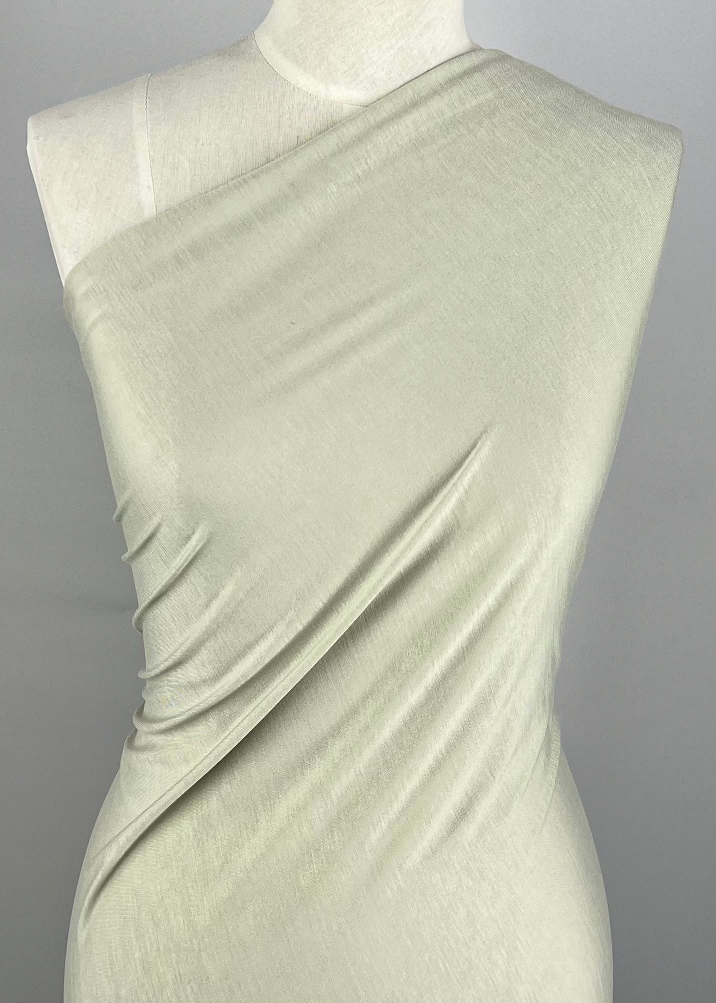 A mannequin stands draped in a pale green, one-shoulder fabric. The light-weight fabric, 100% cotton Cotton Jersey - Alfalfa - 150cm from Super Cheap Fabrics, appears soft and is wrapped snugly around the torso, showcasing a minimalist, elegant style. The plain gray background highlights the fabric and draping technique beautifully.